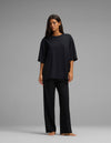 A woman stands barefoot against a gray background, wearing the Cozy Earth Women’s First Layer Boyfriend Sleep Tee & Pajama Pant Set in black, with her hair down and her arms relaxed by her sides.|Color:Jet Black