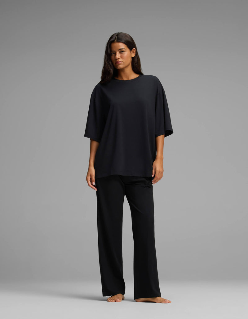 A woman stands barefoot against a gray background, wearing the Cozy Earth Women’s First Layer Boyfriend Sleep Tee & Pajama Pant Set in black, with her hair down and her arms relaxed by her sides.