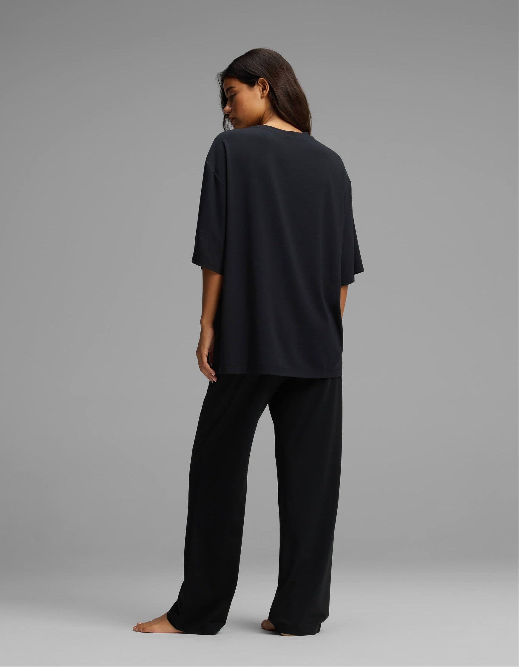 A woman stands barefoot, facing away, wearing the Cozy Earth Women’s First Layer Boyfriend Sleep Tee & Pajama Pant Set in black against a plain gray background.|Color:Jet Black