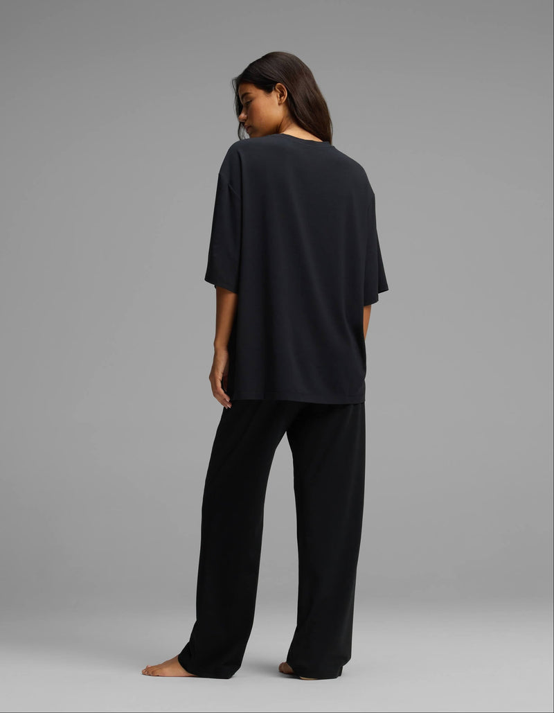 A woman stands barefoot, facing away, wearing the Cozy Earth Women’s First Layer Boyfriend Sleep Tee & Pajama Pant Set in black against a plain gray background.