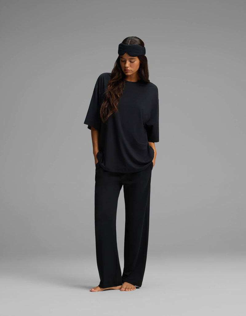 A woman stands barefoot against a gray background, wearing the Cozy Earth Women’s First Layer Boyfriend Sleep Tee & Pajama Pant Set in black. The oversized, relaxed-fit T-shirt and loose pajama pants exude comfort, while a black sleep mask is pushed up on her forehead. Her hands rest in her pockets and her long hair drapes over one shoulder.