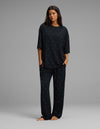 A woman stands barefoot in a relaxed pose, wearing the Cozy Earth Women’s First Layer Boyfriend Sleep Tee & Pajama Pant Set—a loose-fitting black pajama ensemble with small white polka dots—against a plain gray background.|Color:Mini Ghost