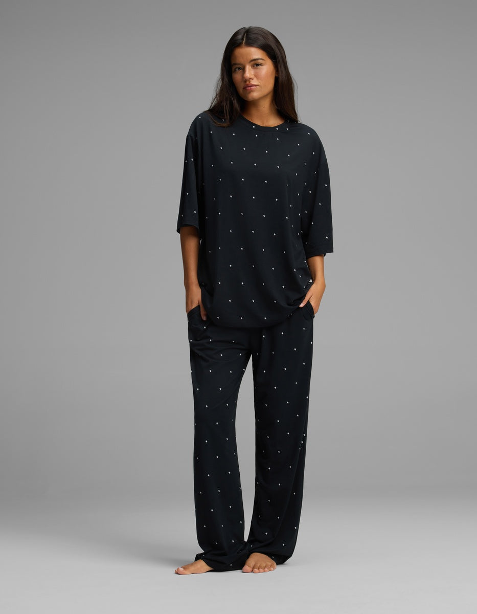 A woman stands barefoot in a relaxed pose, wearing the Cozy Earth Women’s First Layer Boyfriend Sleep Tee & Pajama Pant Set—a loose-fitting black pajama ensemble with small white polka dots—against a plain gray background.|Color:Mini Ghost