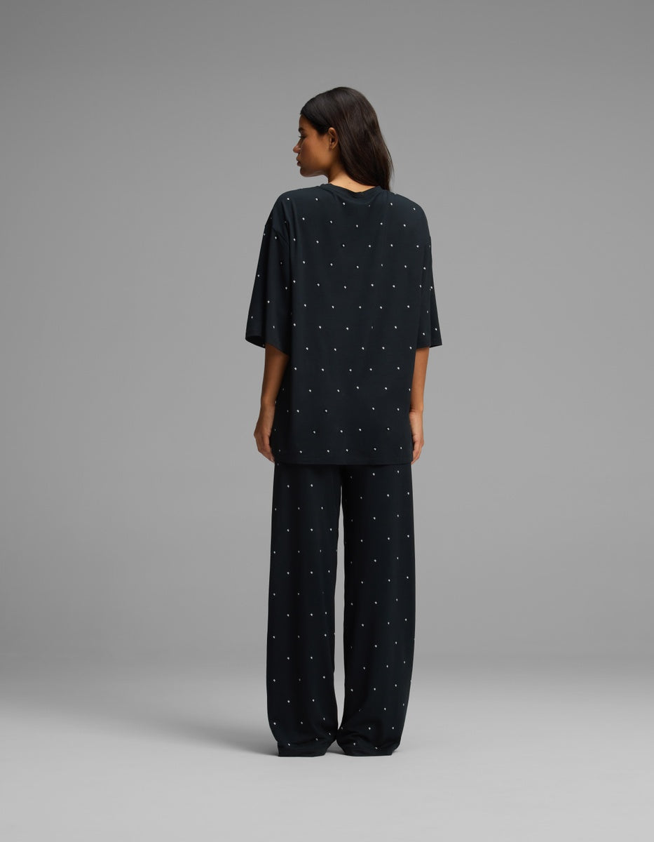 A person stands facing away, wearing the Cozy Earth Women’s First Layer Boyfriend Sleep Tee & Pajama Pant Set in black with small white dot patterns. The background is plain gray, highlighting the set’s relaxed fit.