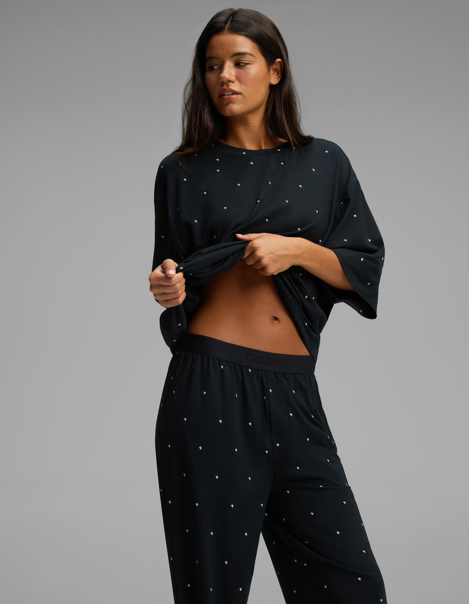 A woman wears the Cozy Earth Women’s First Layer Boyfriend Sleep Tee & Pajama Pant Set in black with small white dots, lifting her top slightly to reveal part of her stomach against a plain gray background.