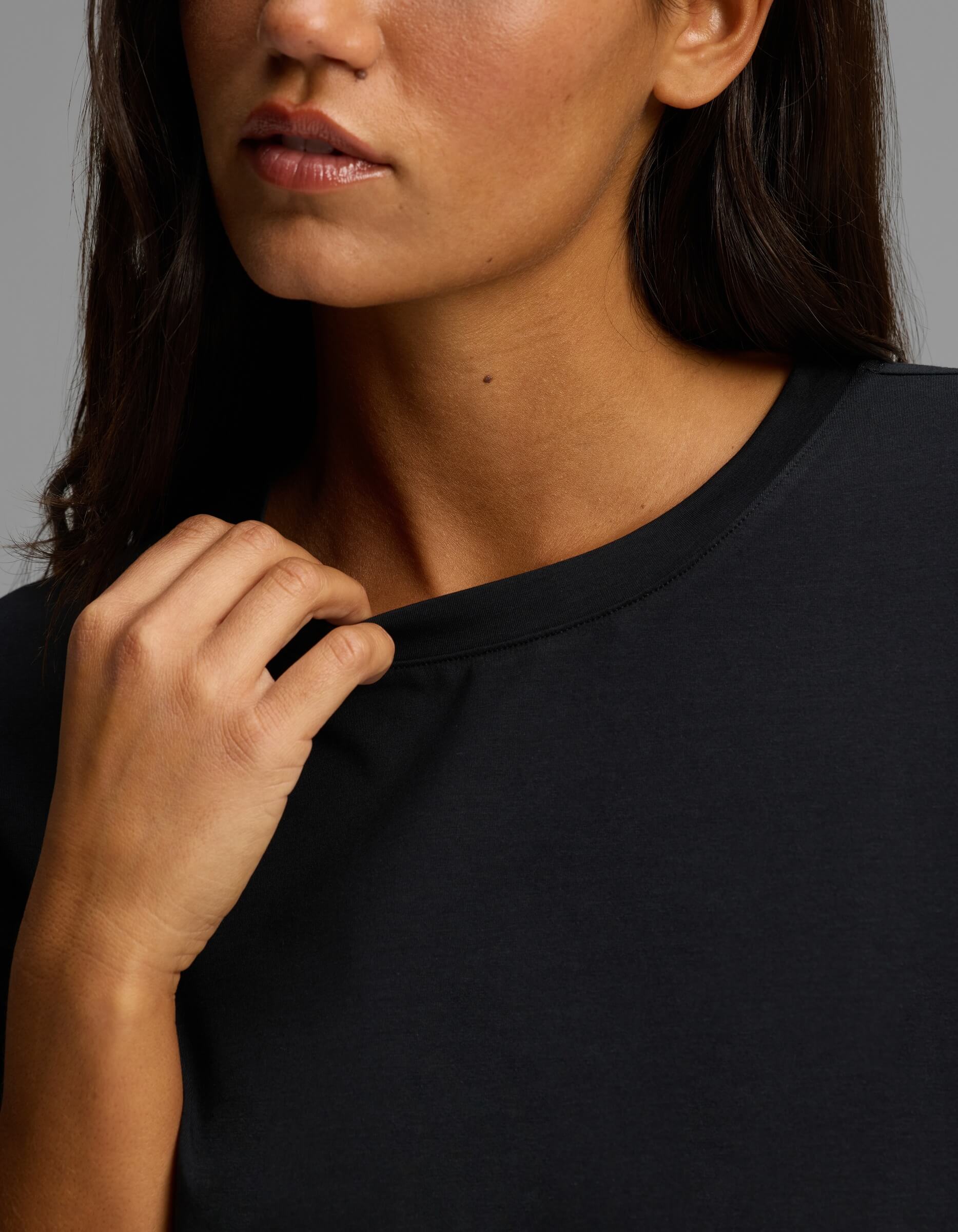 A woman with medium skin tone and straight dark brown hair wears the Cozy Earth Women’s First Layer Boyfriend Sleep Tee in black, gently pulling at its neckline with her right hand. The background is neutral gray, and only the lower half of her face is visible.