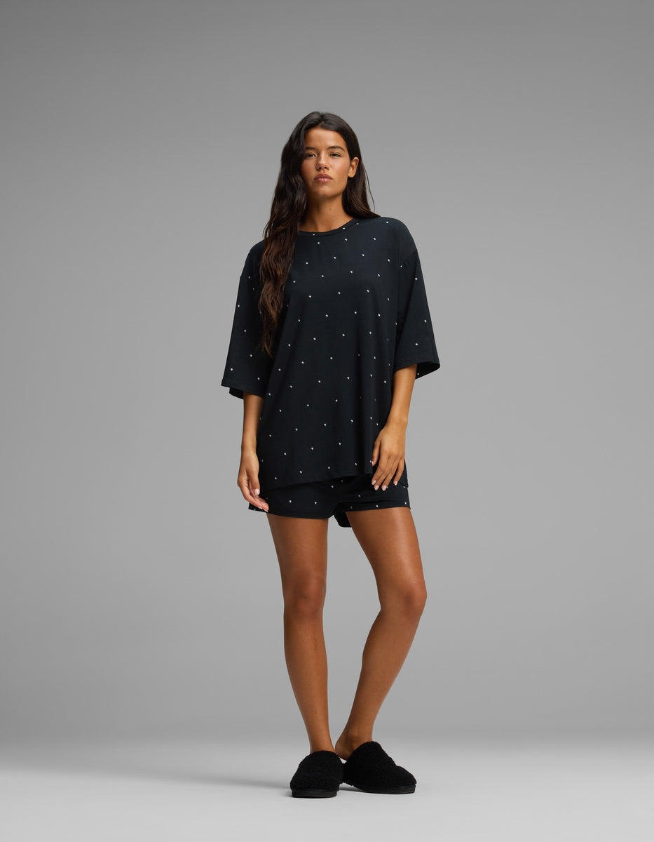 A woman with long brown hair and a neutral expression stands against a gray background wearing the Cozy Earth Women’s First Layer Boyfriend Sleep Tee in black, paired with matching black shorts and black slippers.