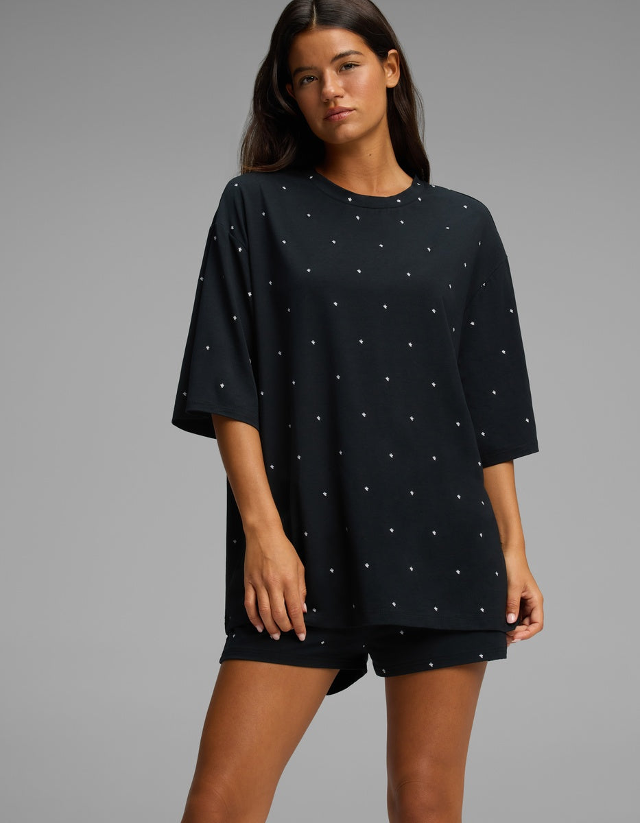 A woman stands against a plain gray background, wearing the Cozy Earth Women’s First Layer Boyfriend Sleep Tee in black with small white polka dots, paired with matching shorts.|Color:Mini Ghost