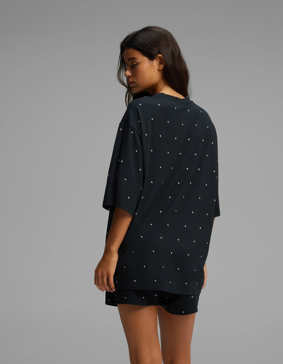 A woman with long dark hair faces away, wearing the Cozy Earth Women’s First Layer Boyfriend Sleep Tee in black, paired with matching shorts featuring small white dots. The background is plain gray.