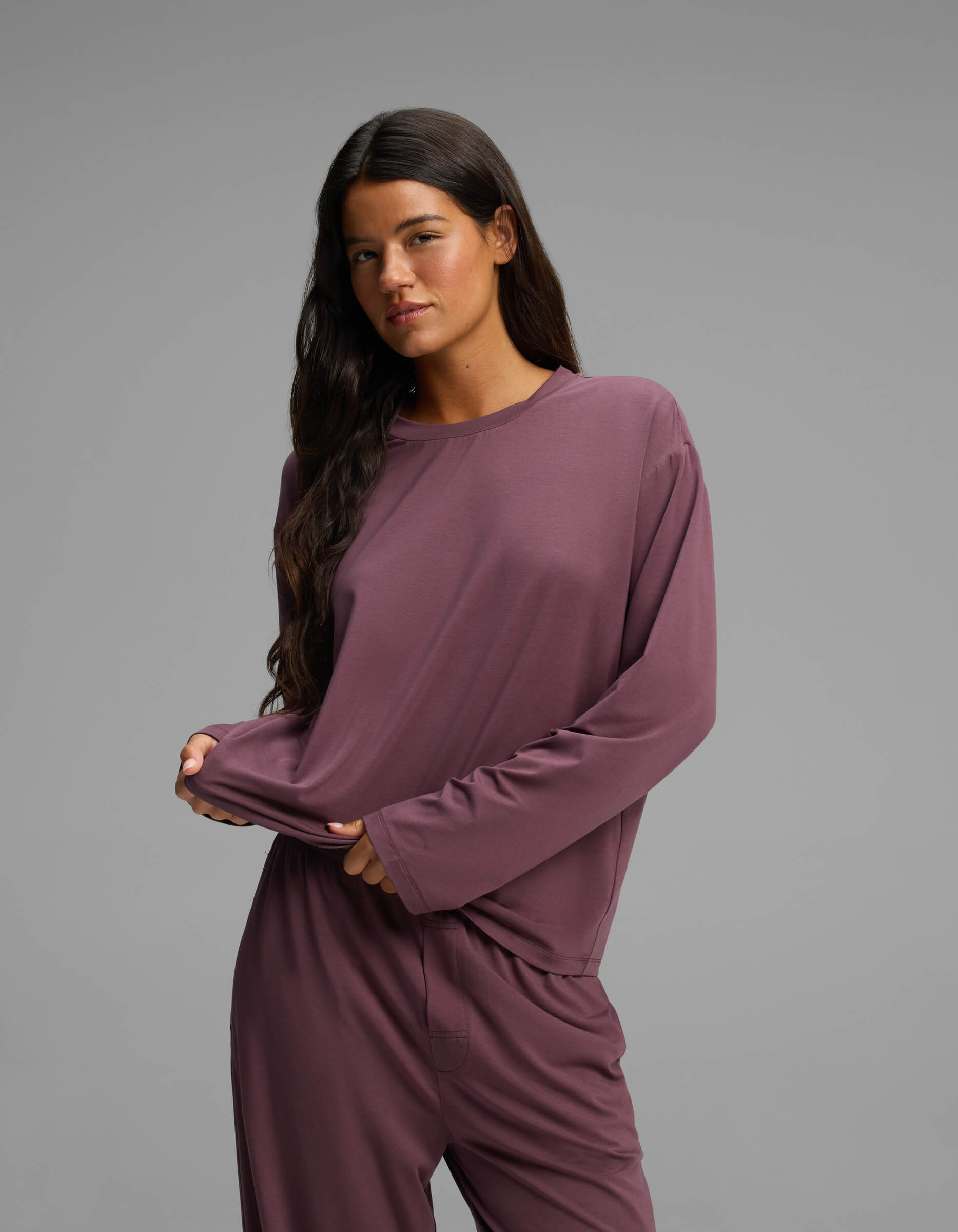 A woman with long dark hair stands against a plain gray background, gently pulling at her shirt with one hand. She is wearing the Cozy Earth Women’s First Layer Long Sleeve Sleep Tee and matching pants in mauve.|Color:Fig