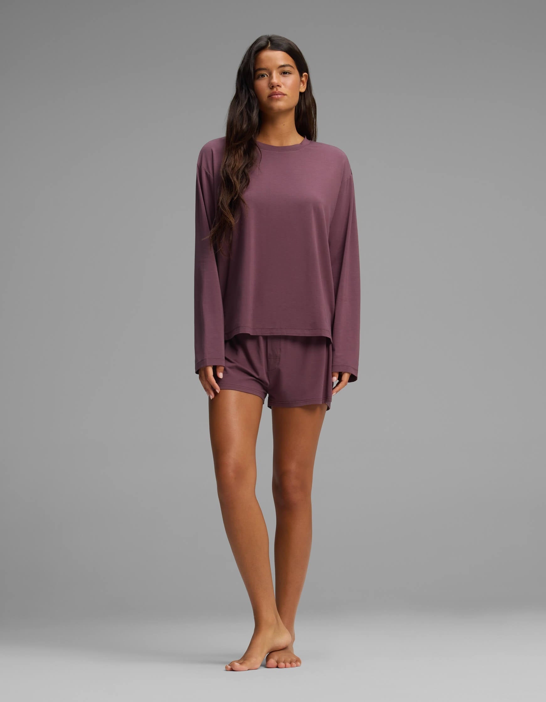 A woman stands barefoot against a plain gray background, wearing the Cozy Earth Women’s First Layer Long Sleeve Sleep Tee & Boxer Short Set in mauve, her long dark hair draped over her shoulder.