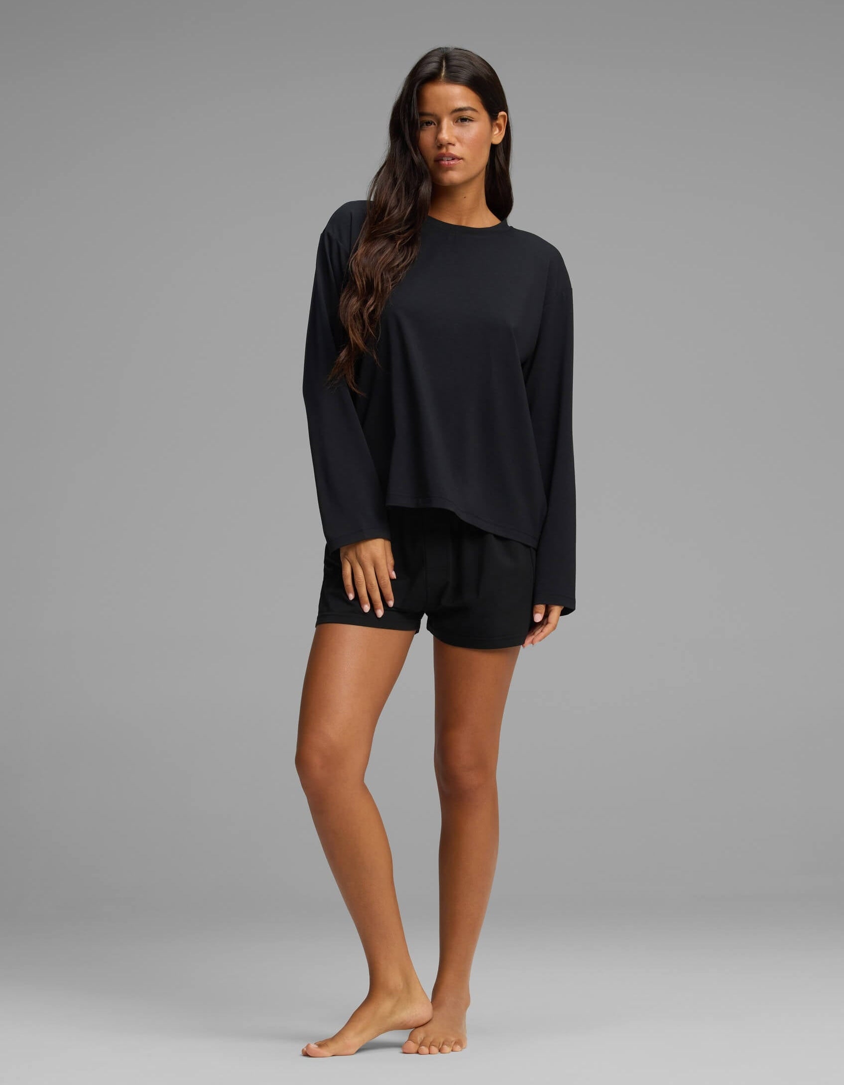 A woman with long brown hair stands barefoot on a grey background, modeling the Cozy Earth Women’s First Layer Long Sleeve Sleep Tee & Boxer Short Set in black, looking at the camera with a neutral expression.|Color:Jet Black