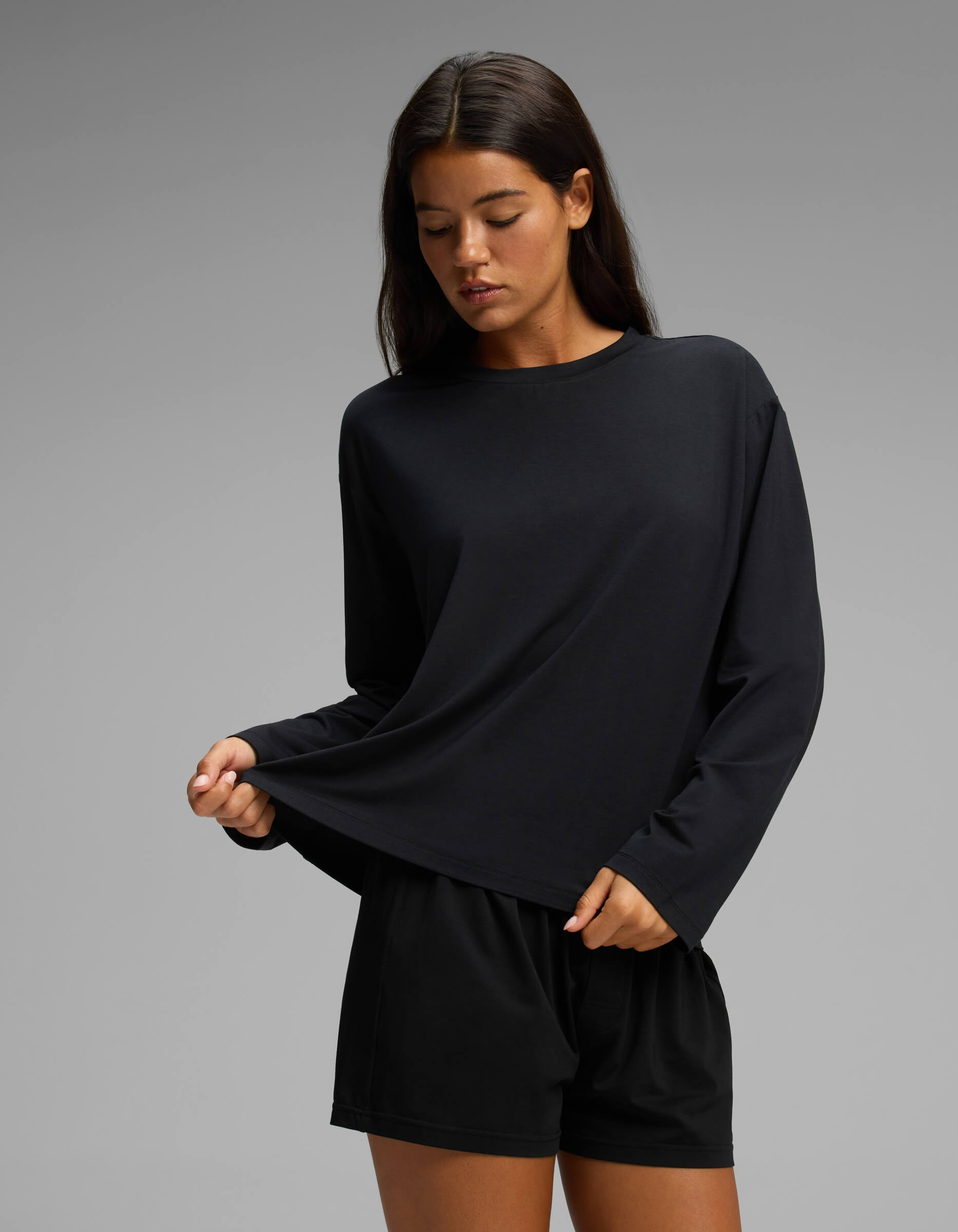 A woman stands against a plain gray background, gently pulling at the hem of her black Cozy Earth Women’s First Layer Long Sleeve Sleep Tee & Boxer Short Set while looking down.|Color:Jet Black