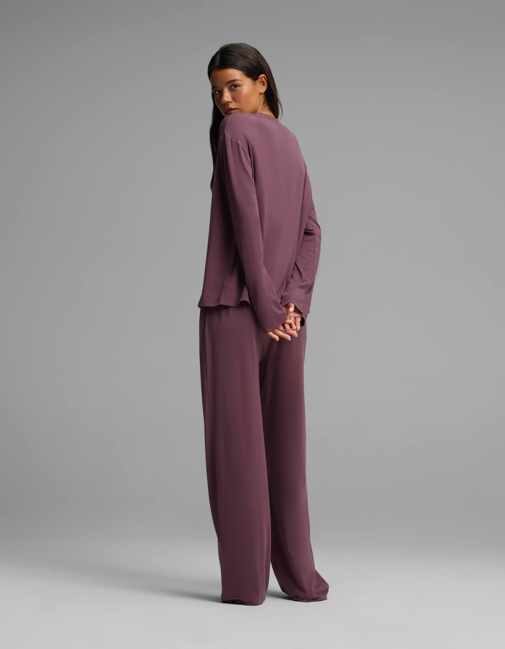 A woman stands with her back partially turned against a plain gray background, her hands clasped behind her. She is wearing the Cozy Earth Women’s First Layer Long Sleeve Sleep Tee & Pajama Pant Set in mauve, featuring a loose fit and wide-leg silhouette.|Color:Fig