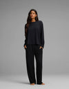A woman with long brown hair stands facing forward against a plain gray background. She is wearing the Cozy Earth Women’s First Layer Long Sleeve Sleep Tee & Pajama Pant Set in black, with her hands tucked into the side pockets. She is barefoot.|Color:Jet Black