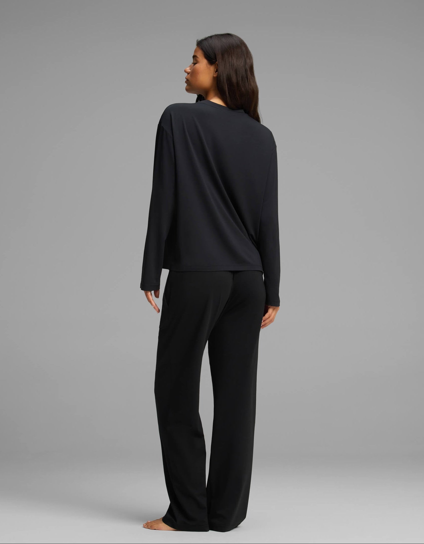 A woman with long brown hair, seen from behind, stands barefoot against a plain gray background wearing the Cozy Earth Women’s First Layer Long Sleeve Sleep Tee & Pajama Pant Set in black.|Color:Jet Black
