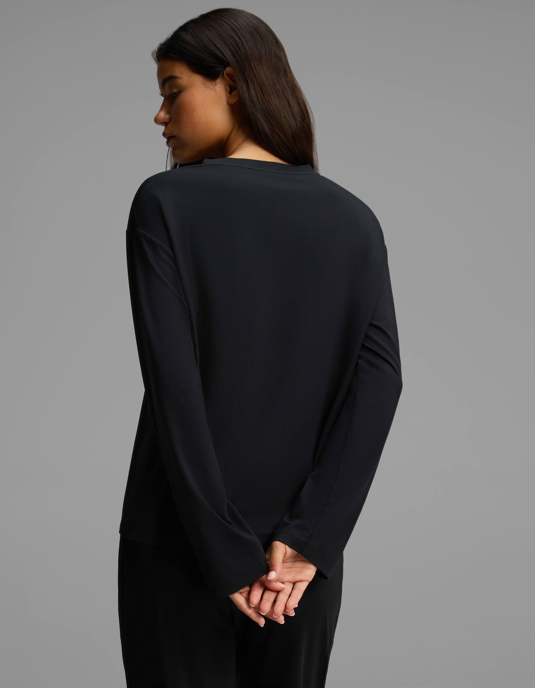 A woman with long dark hair stands facing away, her head turned to the side. She is wearing Cozy Earth’s Women’s First Layer Long Sleeve Sleep Tee in black, paired with black pants, her hands clasped behind her back against a plain gray background.|Color:Jet Black