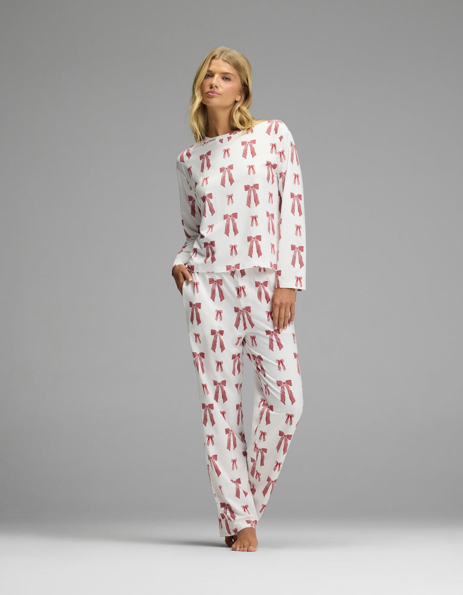 A woman wearing a Cozy Earth Women’s First Layer Long Sleeve Sleep Tee & Pajama Pant Set in white with a red bow pattern.
