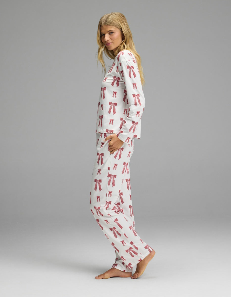 A woman wearing a white long-sleeved Cozy Earth Women’s First Layer Long Sleeve Sleep Tee & Pajama Pant Set featuring an all-over pattern of red bows.