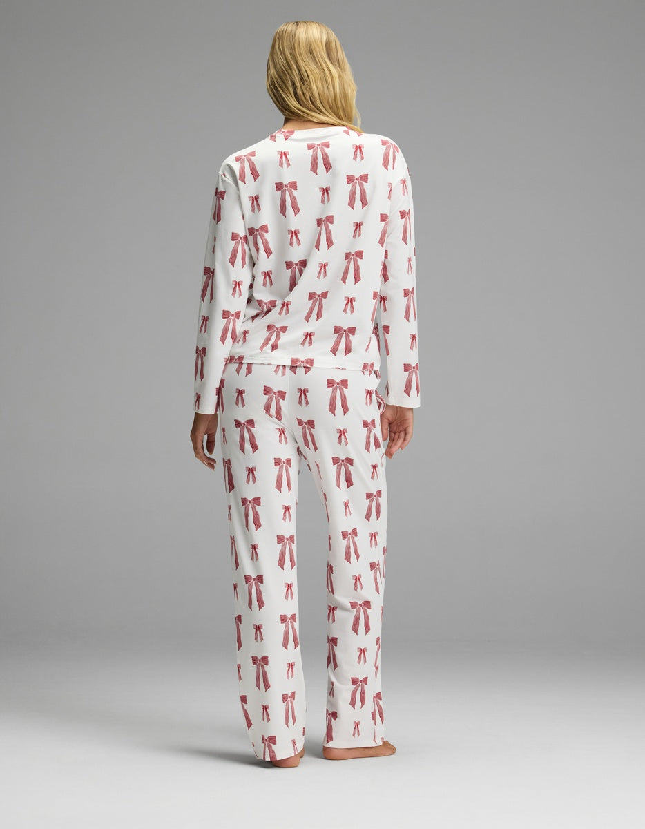 A woman wearing the Cozy Earth Women’s First Layer Long Sleeve Sleep Tee & Pajama Pant Set stands with her back to the camera, showcasing the white long-sleeve pajamas with an all-over red bow print.