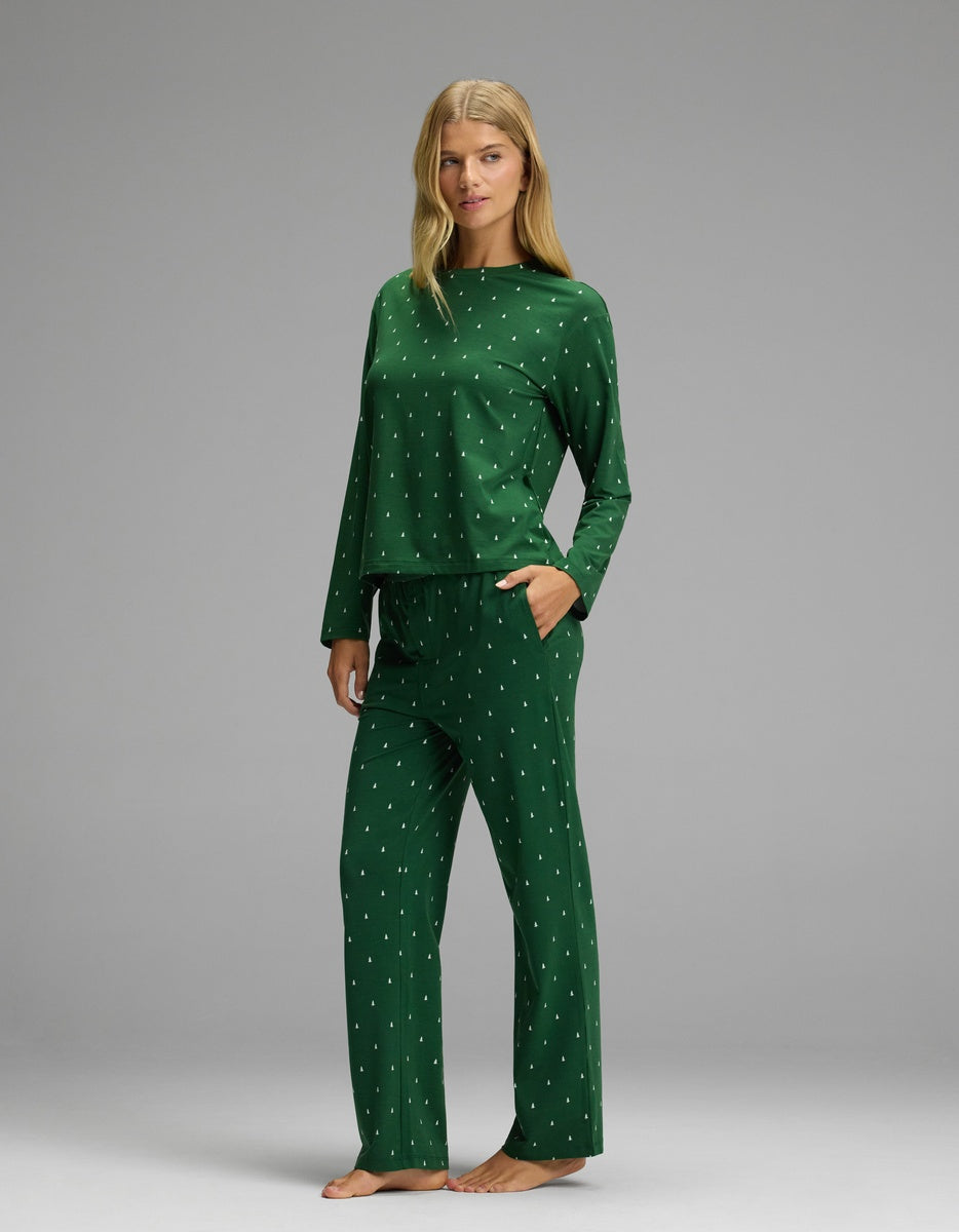 A woman wearing a green Cozy Earth Women’s First Layer Long Sleeve Sleep Tee & Pajama Pant Set featuring a small white tree print.