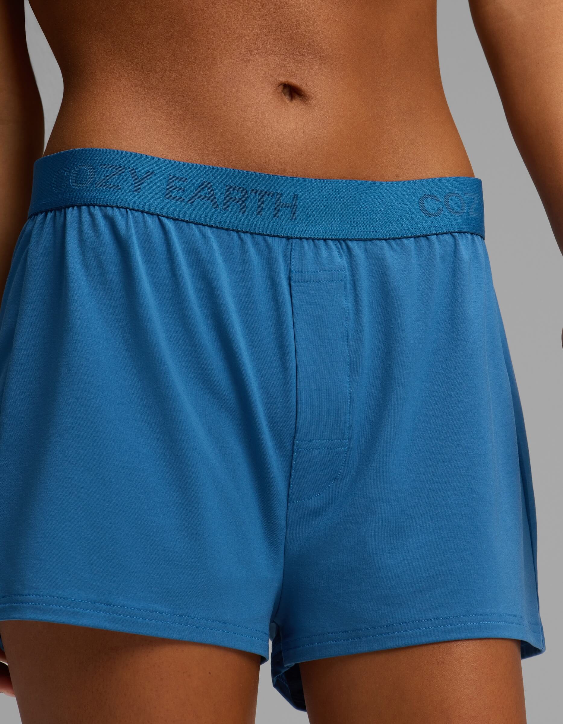 A person wearing Cozy Earth Women’s First Layer Pajama Boxer Shorts, featuring the brand name on the waistband, stands against a plain background.