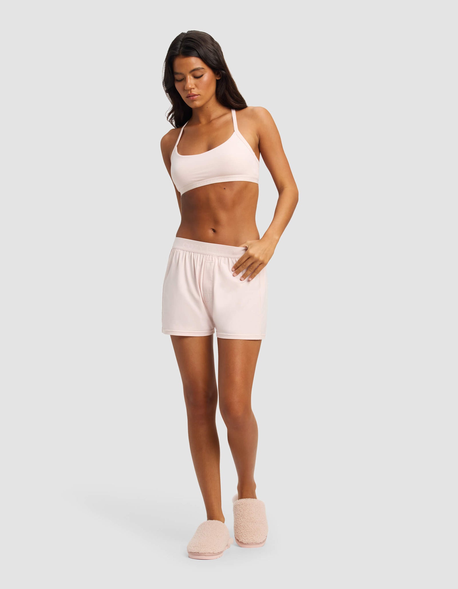 A woman stands against a light gray background wearing Cozy Earth’s Women's First Layer Pajama Boxer Short in light pink, with a matching bralette and slippers. She looks down, one hand on her shorts and the other at her side.