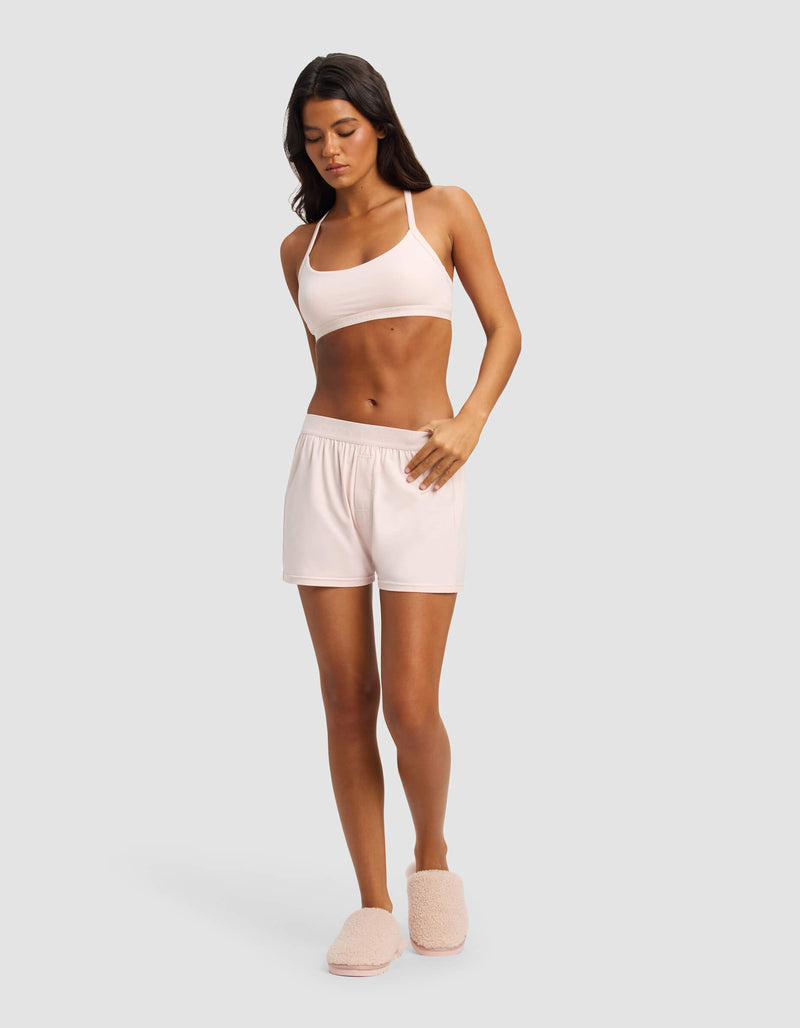 A woman stands against a light gray background wearing Cozy Earth’s Women's First Layer Pajama Boxer Short in light pink, with a matching bralette and slippers. She looks down, one hand on her shorts and the other at her side.