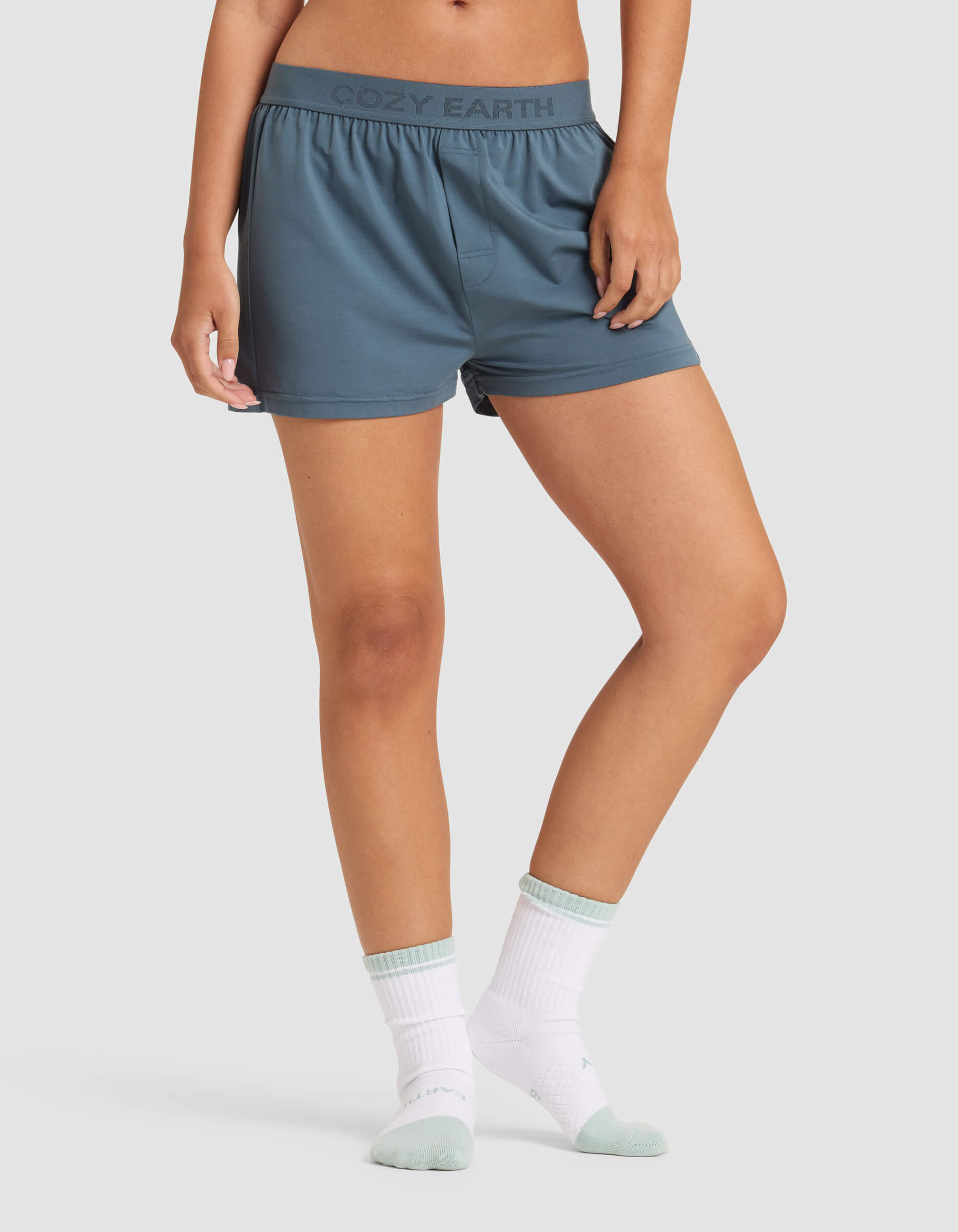 A person is shown from the waist down wearing Cozy Earth Women's First Layer Pajama Boxer Shorts in blue-gray, featuring the brand logo on the waistband, paired with white socks and a white top, set against a plain light background.