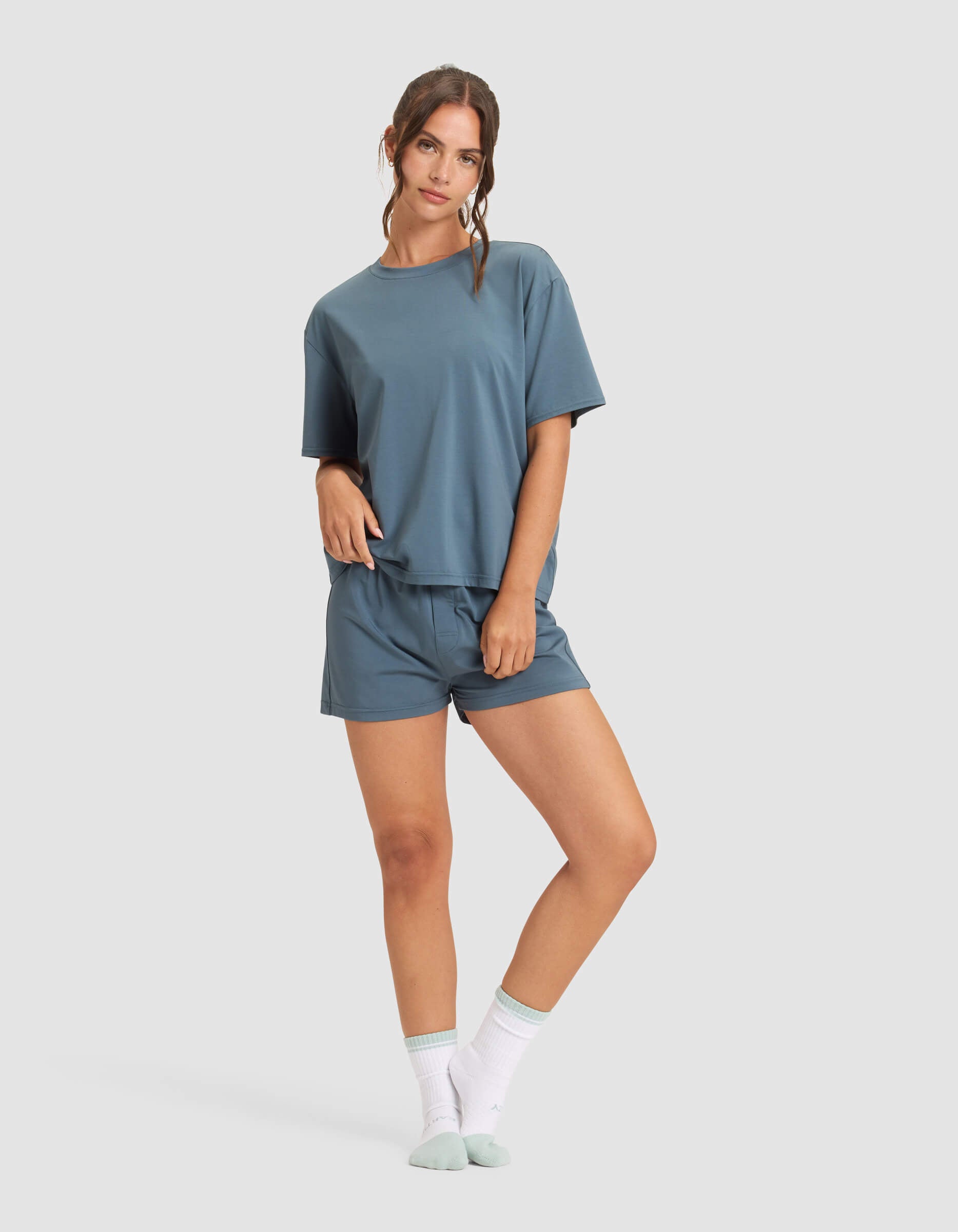 A woman poses casually in the Cozy Earth Women's First Layer Pajama Short Sleeve Tee & Boxer Short Set, featuring a loose fit in blue, styled with white crew socks and light blue slippers against a plain background.|Color:Dawn