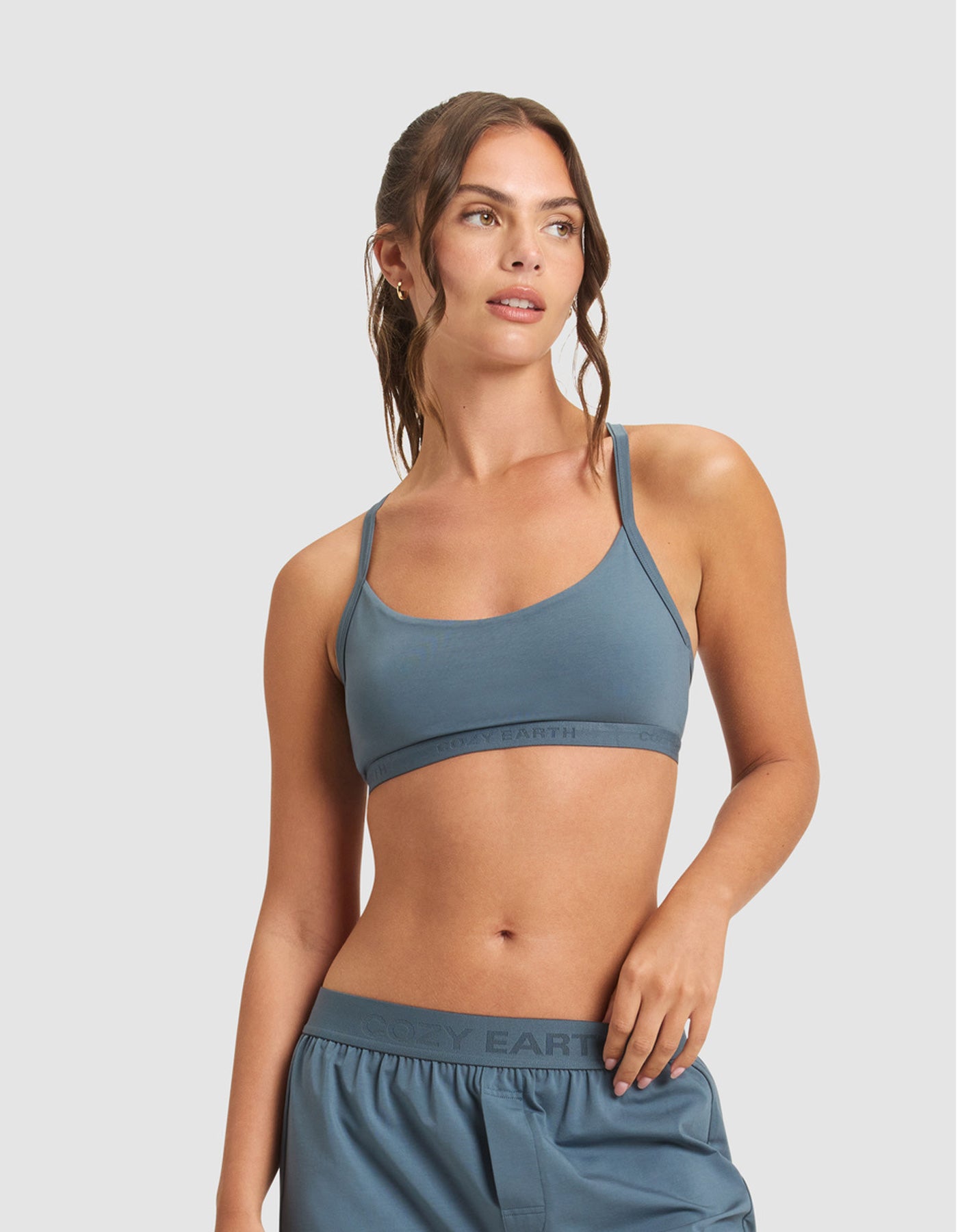 A woman with wavy brown hair in a ponytail wears the Cozy Earth Women's First Layer Pajama Soft Bra and matching shorts, looking to the side against a plain light gray background.
