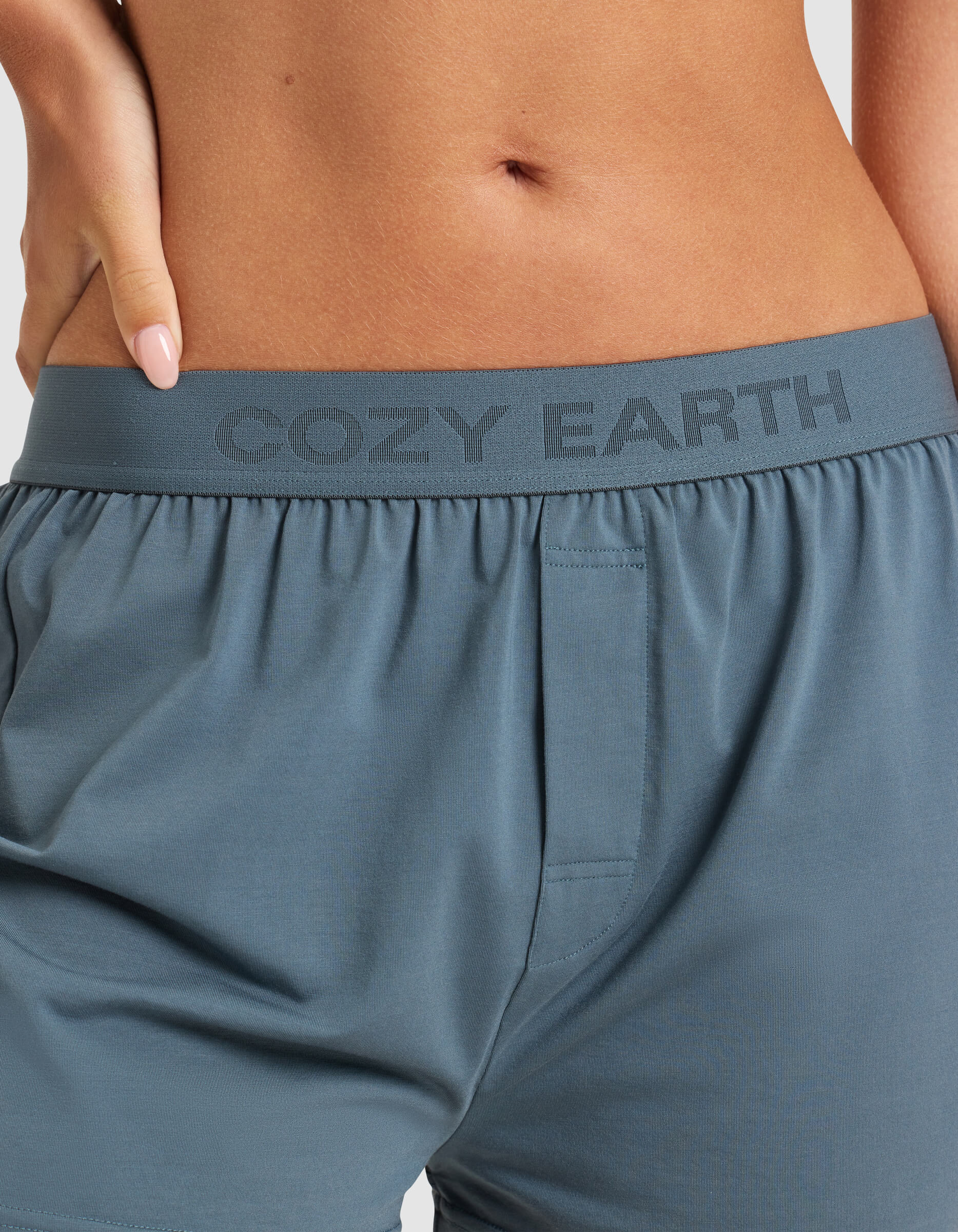 A close-up shows a person wearing Cozy Earth Women’s First Layer Pajama Boxer Shorts in blue-gray, with the branded waistband visible and their hand resting on their hip.