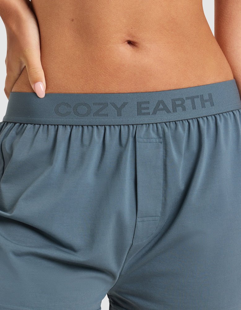 Close-up of a person wearing Cozy Earth's Women's First Layer Pajama Soft Bra & Boxer Short Set in blue-gray, with "COZY EARTH" on the waistband. Their hand rests on their hip, showing part of their midriff and painted nails.