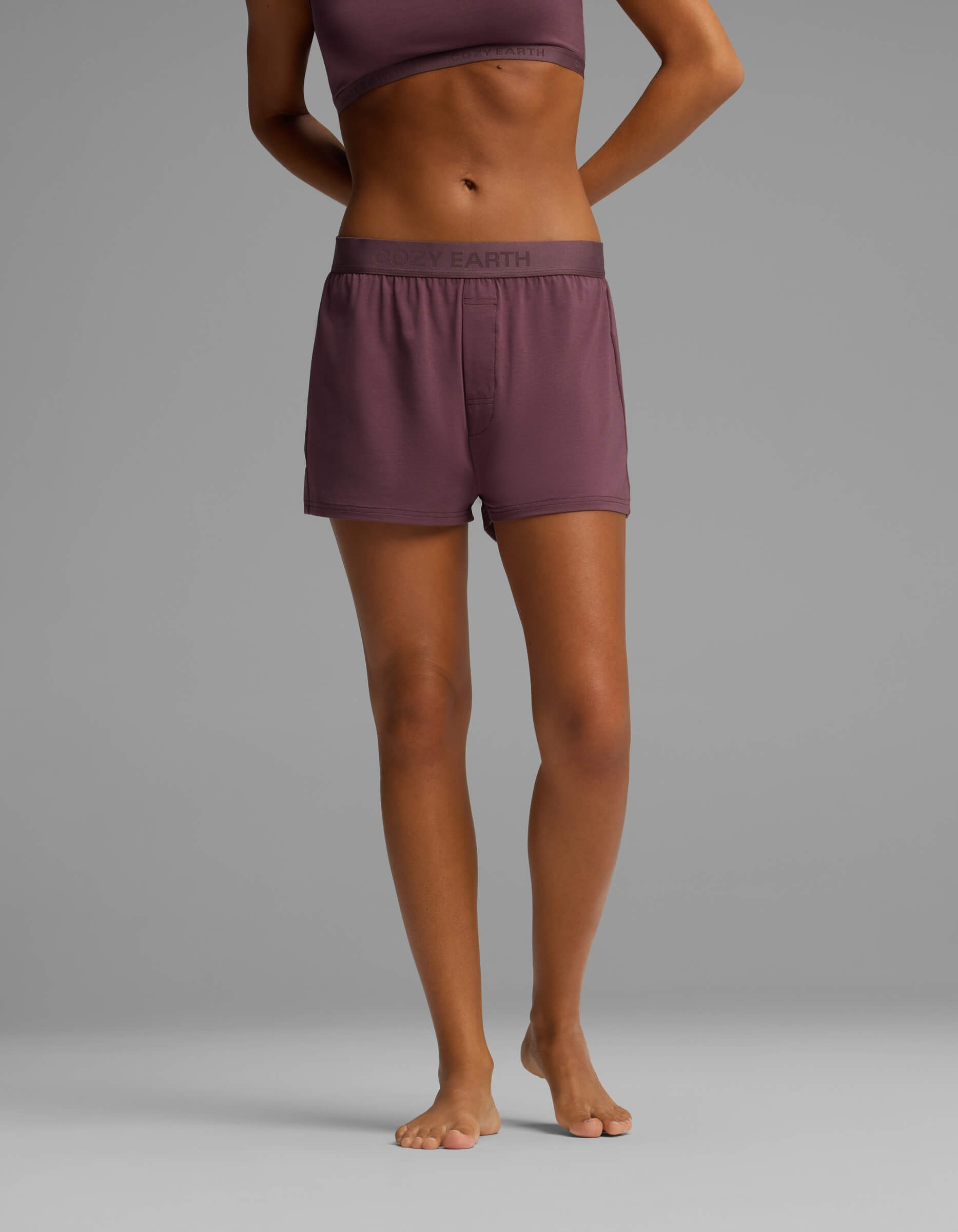 A person standing barefoot against a plain gray background, wearing Cozy Earth’s Women’s First Layer Pajama Boxer Short in mauve with a matching sports bra. Only the body from shoulders to feet is visible.|Color:Fig