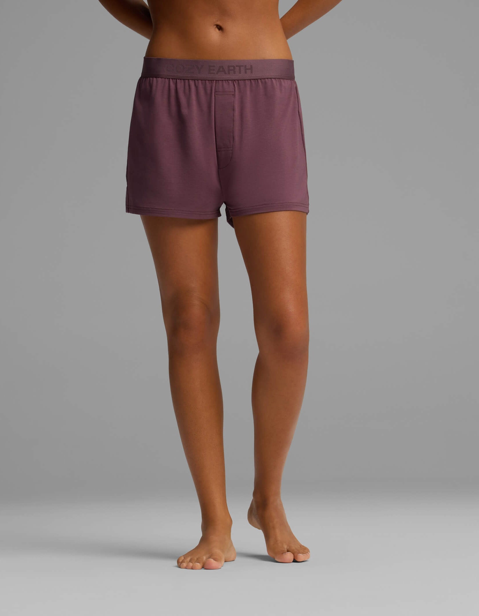 A person stands barefoot on a gray background, wearing mauve boxer shorts from the Cozy Earth Women’s First Layer Pajama Short Sleeve Tee & Boxer Short Set, featuring a waistband with the brand name “COZY EARTH.” Only the lower torso and legs are visible.