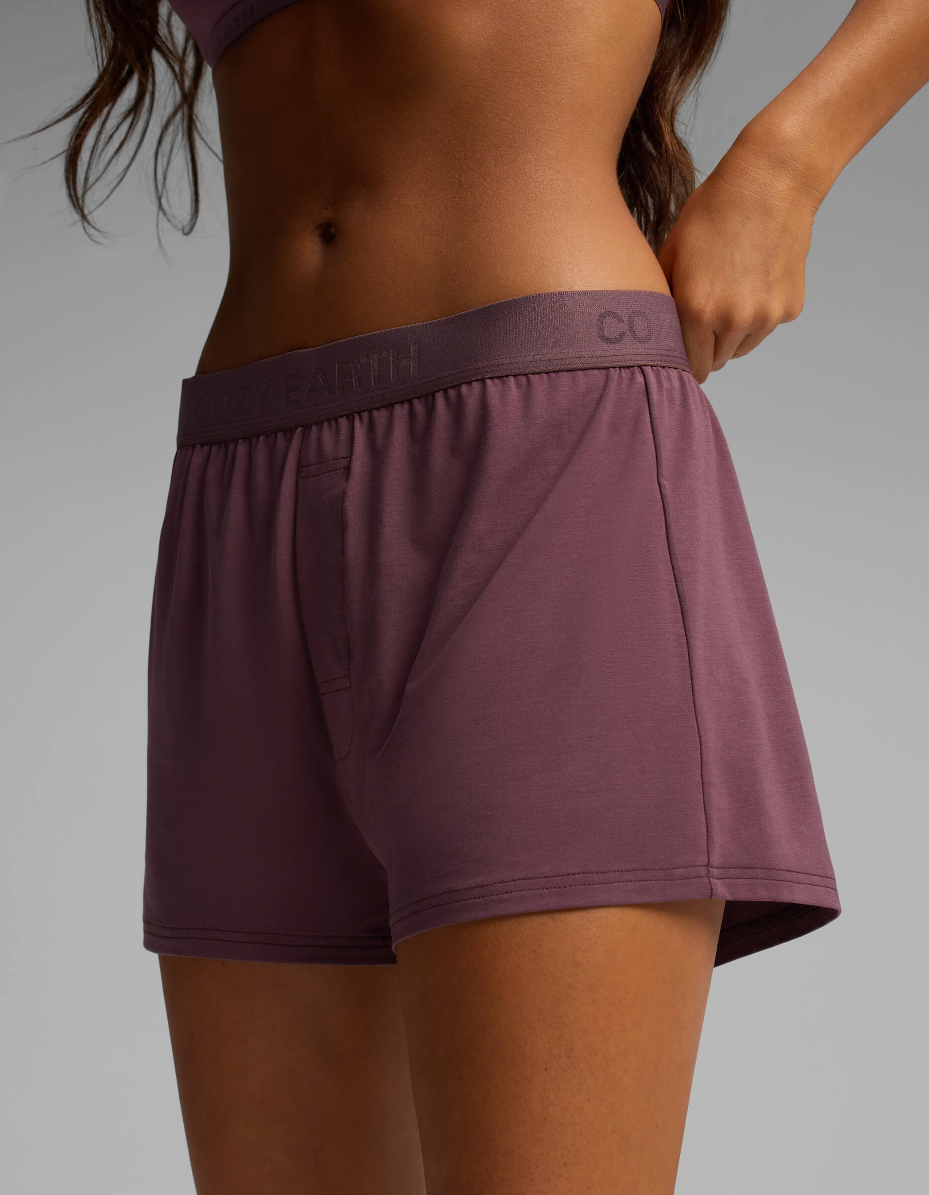 A person is shown from the waist to mid-thigh wearing mauve-colored Cozy Earth Women’s First Layer Pajama Boxer Shorts, with one hand resting on the waistband against a plain gray background.