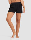 A woman is shown from torso to legs, barefoot against a plain light gray background, wearing Cozy Earth's Women's First Layer Pajama Boxer Short in black.|Color:Jet Black
