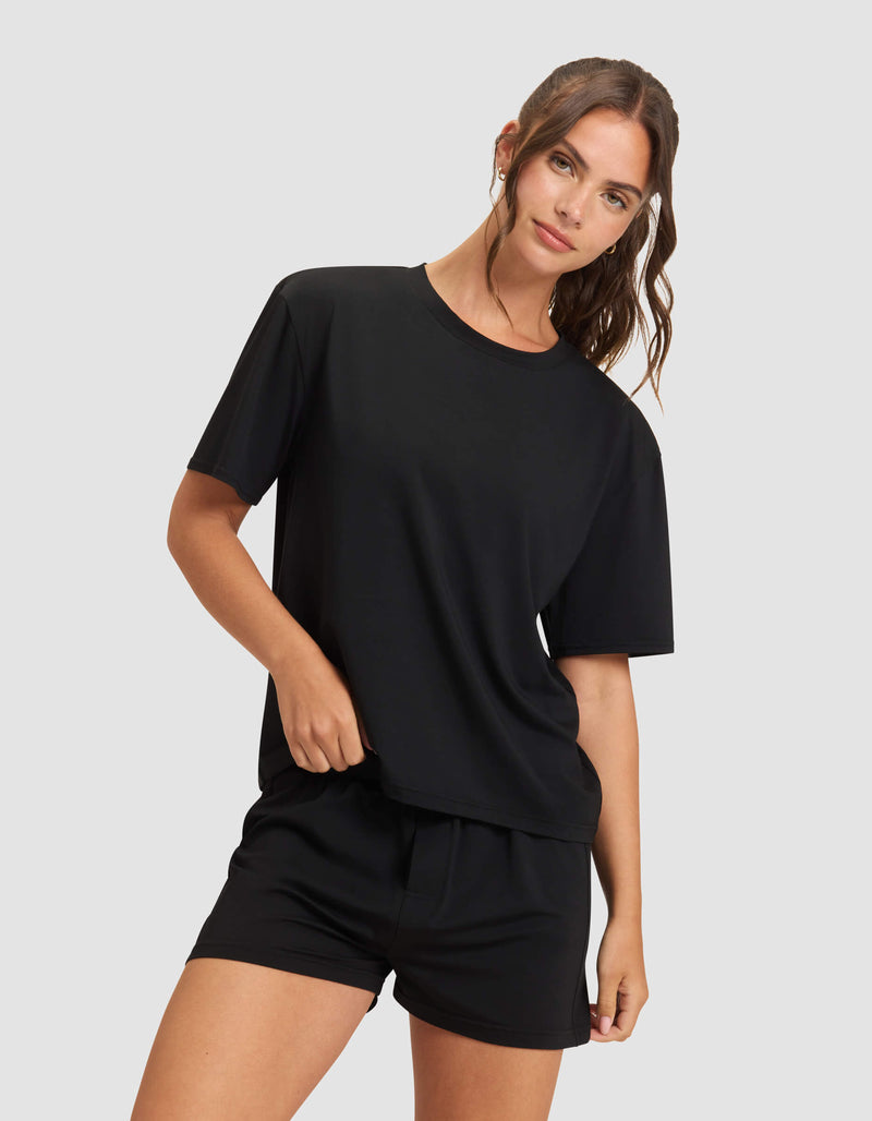 A woman with long brown hair tied back models the Cozy Earth Women's First Layer Pajama Boxer Short, paired with a loose black T-shirt, standing against a plain light gray background with one hand on her hip and a relaxed expression.