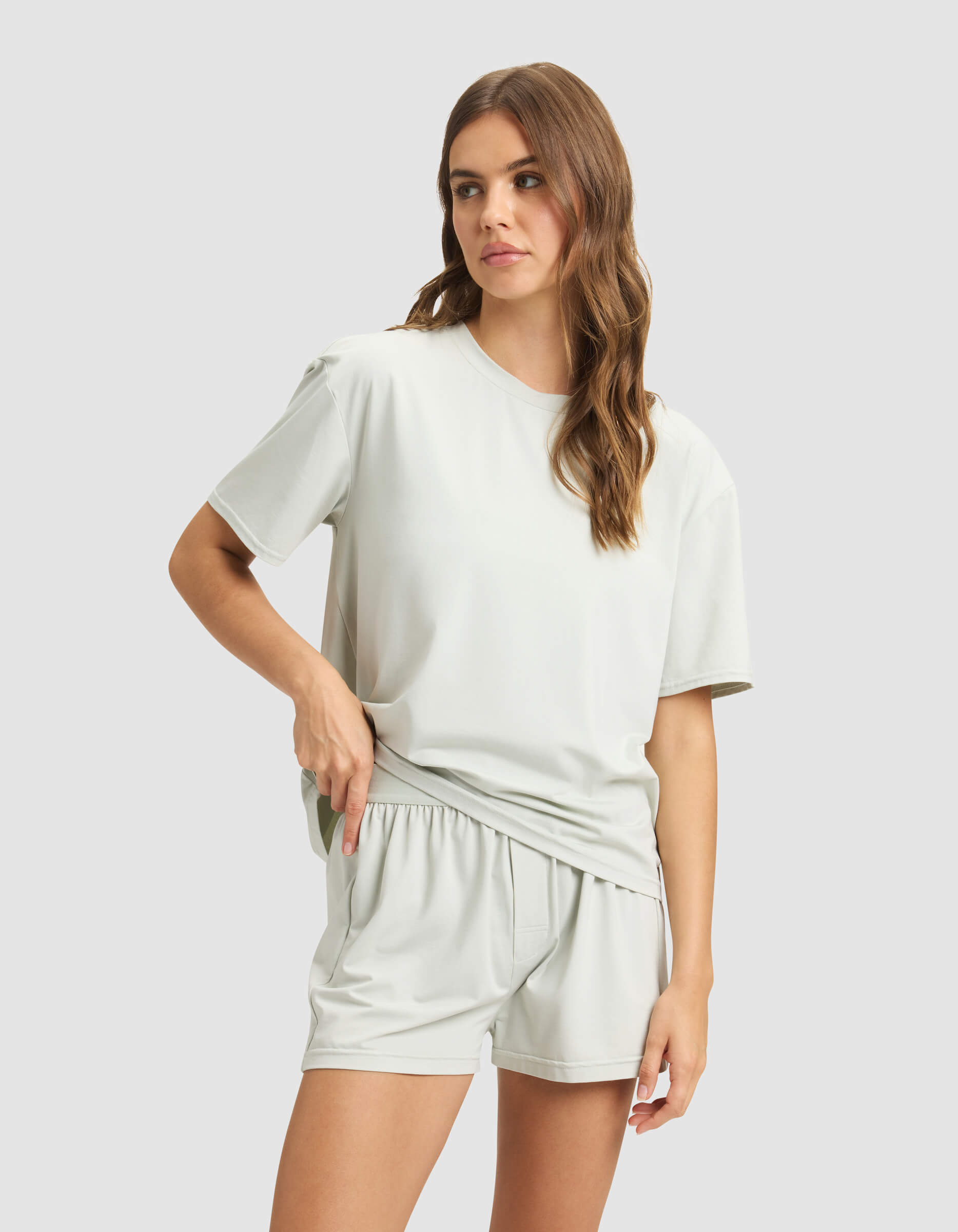 A woman with long brown hair models Cozy Earth’s Women's First Layer Pajama Boxer Short and a loose light gray T-shirt, standing against a plain background with one hand on her hip.|Color:Laurel