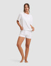 A woman stands barefoot against a plain background, wearing the Cozy Earth Women's First Layer Pajama Short Sleeve Tee & Boxer Short Set. She holds the hem of her shirt and looks ahead with a relaxed expression.|Color:White