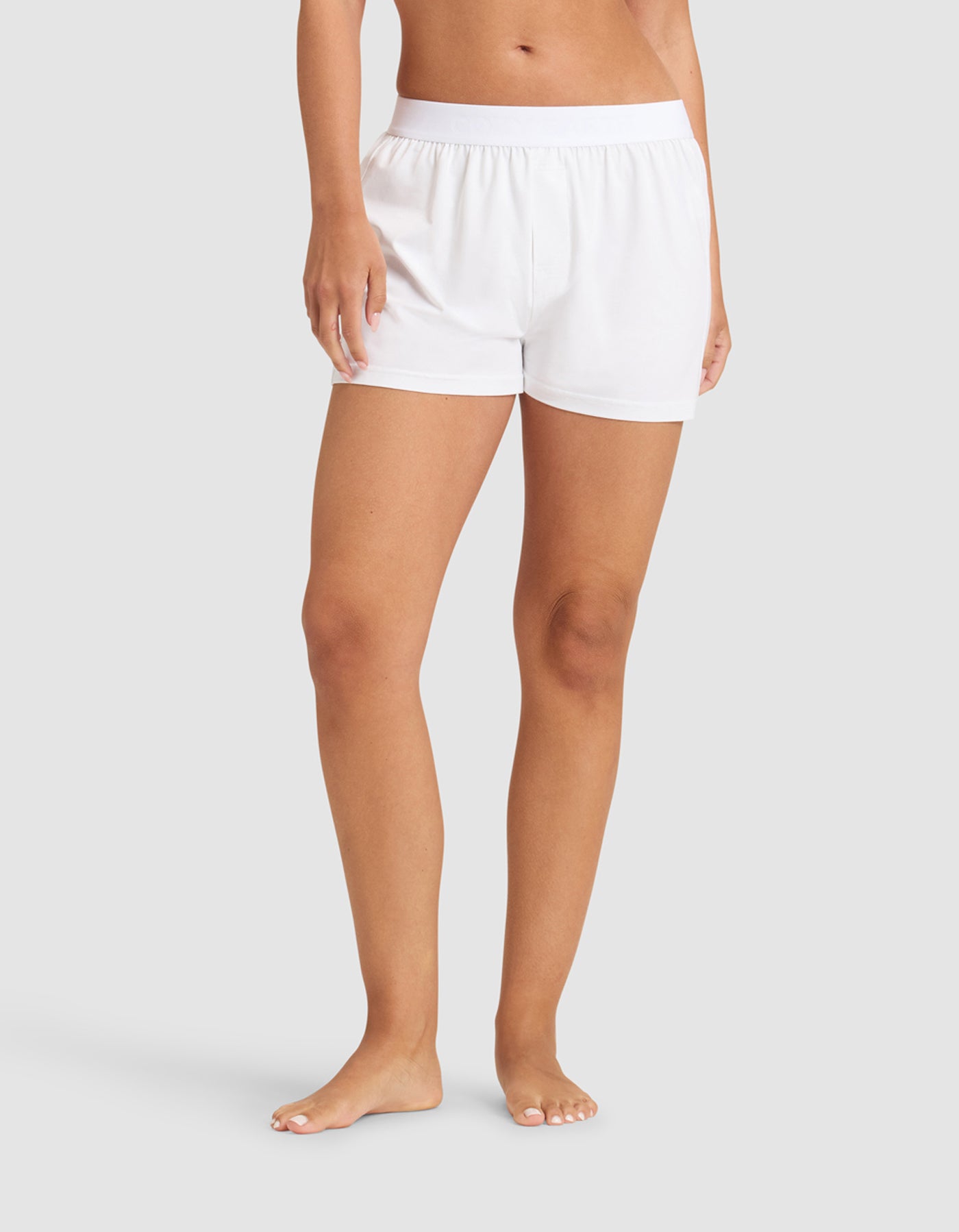 A person stands barefoot against a plain background, wearing Cozy Earth's Women's First Layer Pajama Boxer Short in white with a matching top, arms relaxed by their sides.|Color:White