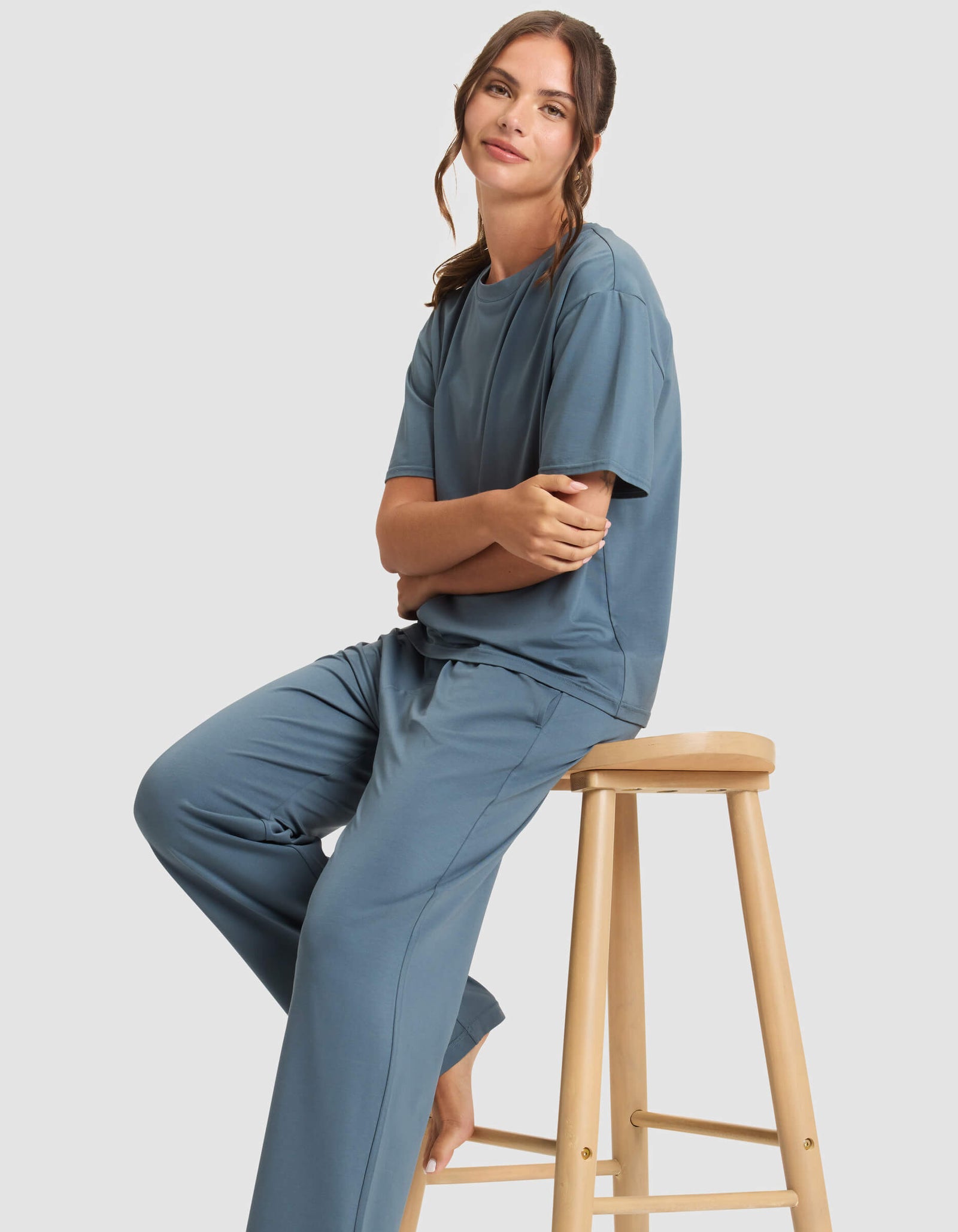 A woman in Cozy Earth's Women's First Layer Pajama Short Sleeve Tee & Pant Set sits barefoot on a wooden stool, arms folded and smiling slightly, against a plain light gray background.
