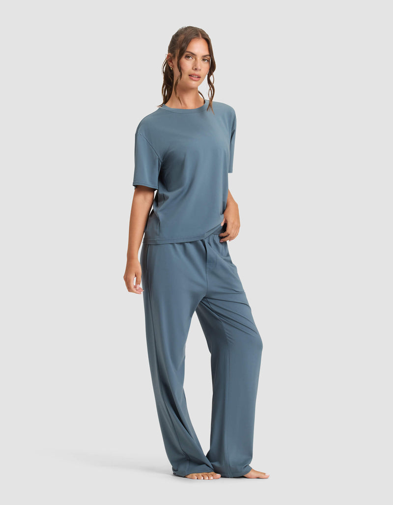A woman stands barefoot against a plain background, wearing the Cozy Earth Women's First Layer Pajama Short Sleeve Tee & Pant Set. Her wavy hair is tied back, and she has a relaxed expression.