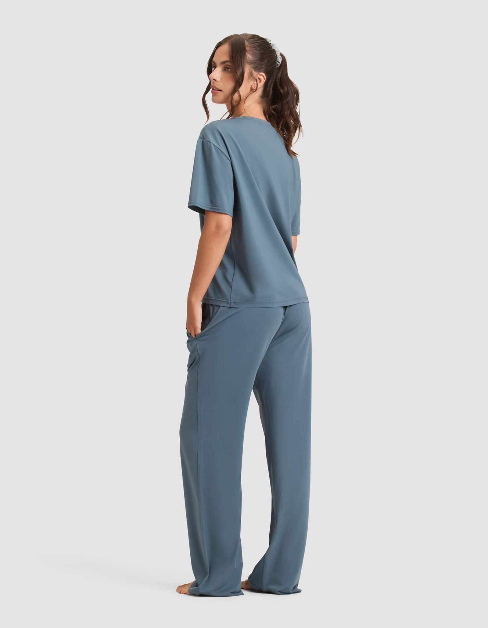 A woman stands barefoot, facing away, wearing the Cozy Earth Women's First Layer Pajama Short Sleeve Tee & Pant Set in blue. She has long brown hair in a ponytail against a plain white background.