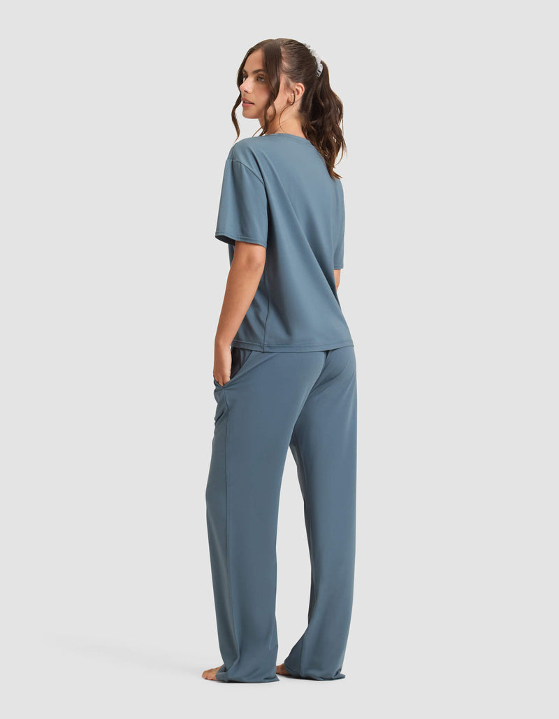 A woman stands barefoot, facing away, wearing the Cozy Earth Women's First Layer Pajama Short Sleeve Tee & Pant Set in blue. She has long brown hair in a ponytail against a plain white background.