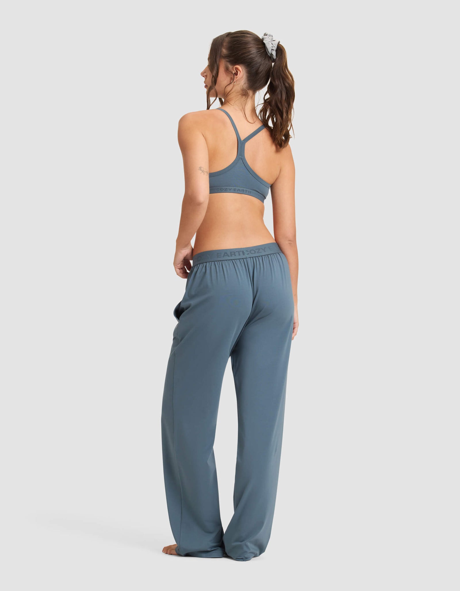A woman, barefoot and with her hair in a scrunchie, models the Cozy Earth Women's First Layer Pajama Soft Bra & Pant Set in blue, standing against a plain light background and glancing over her shoulder.