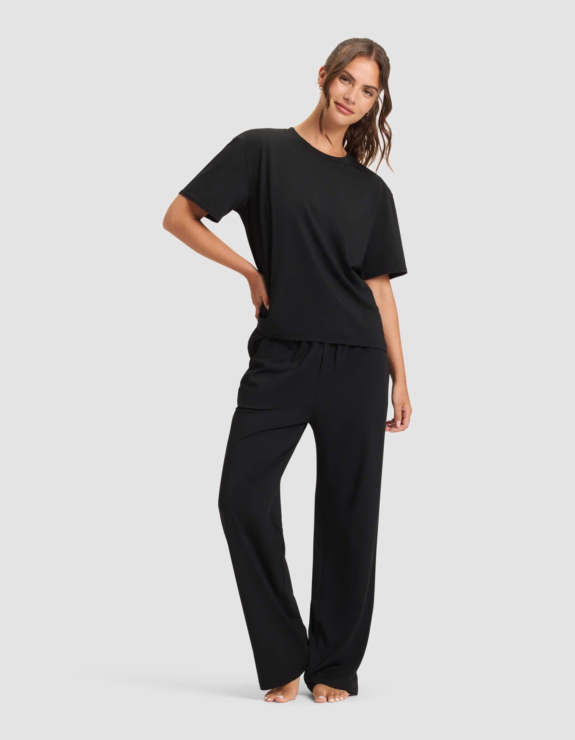 Woman barefoot against a plain background, wearing Cozy Earth Women's First Layer Pajama Short Sleeve Tee & Pant Set in black, with one hand in her pocket and the other by her side.