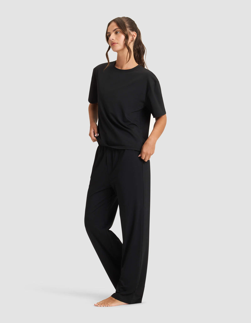 A woman stands barefoot against a plain background, wearing the Cozy Earth Women's First Layer Pajama Short Sleeve Tee & Pant Set in black. She has long brown hair in loose waves and looks slightly to the side.