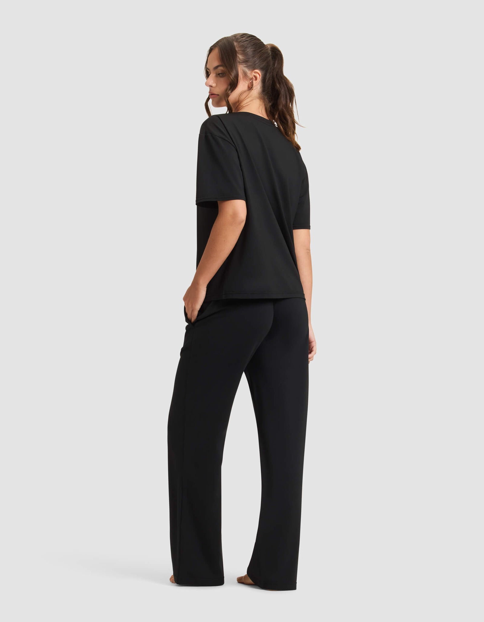 A woman with long brown hair in a ponytail stands barefoot against a light background, wearing the Cozy Earth Women's First Layer Pajama Short Sleeve Tee & Pant Set in black.