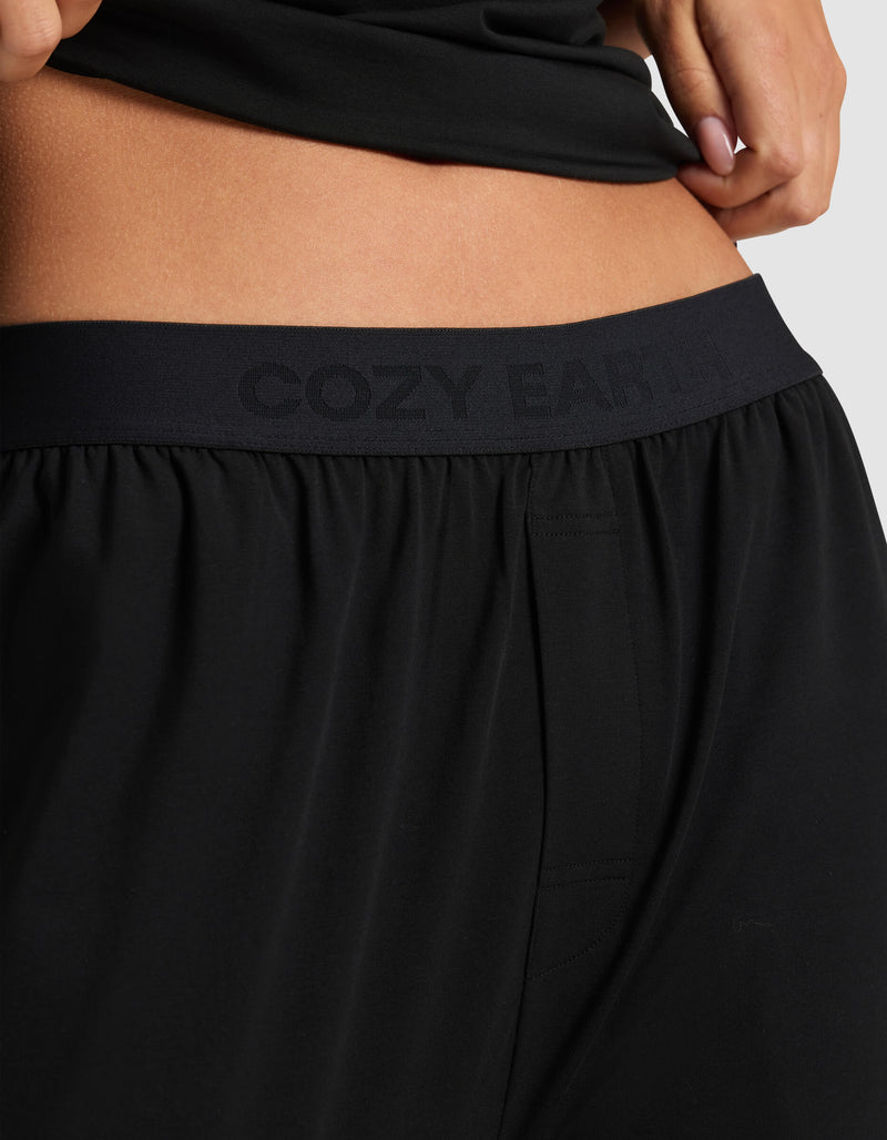 Close-up of a person wearing Cozy Earth Women's First Layer Pajama Pant in black, featuring a "COZY EARTH" waistband. The person's hand rests near their hip, with part of a black top visible above the pants.